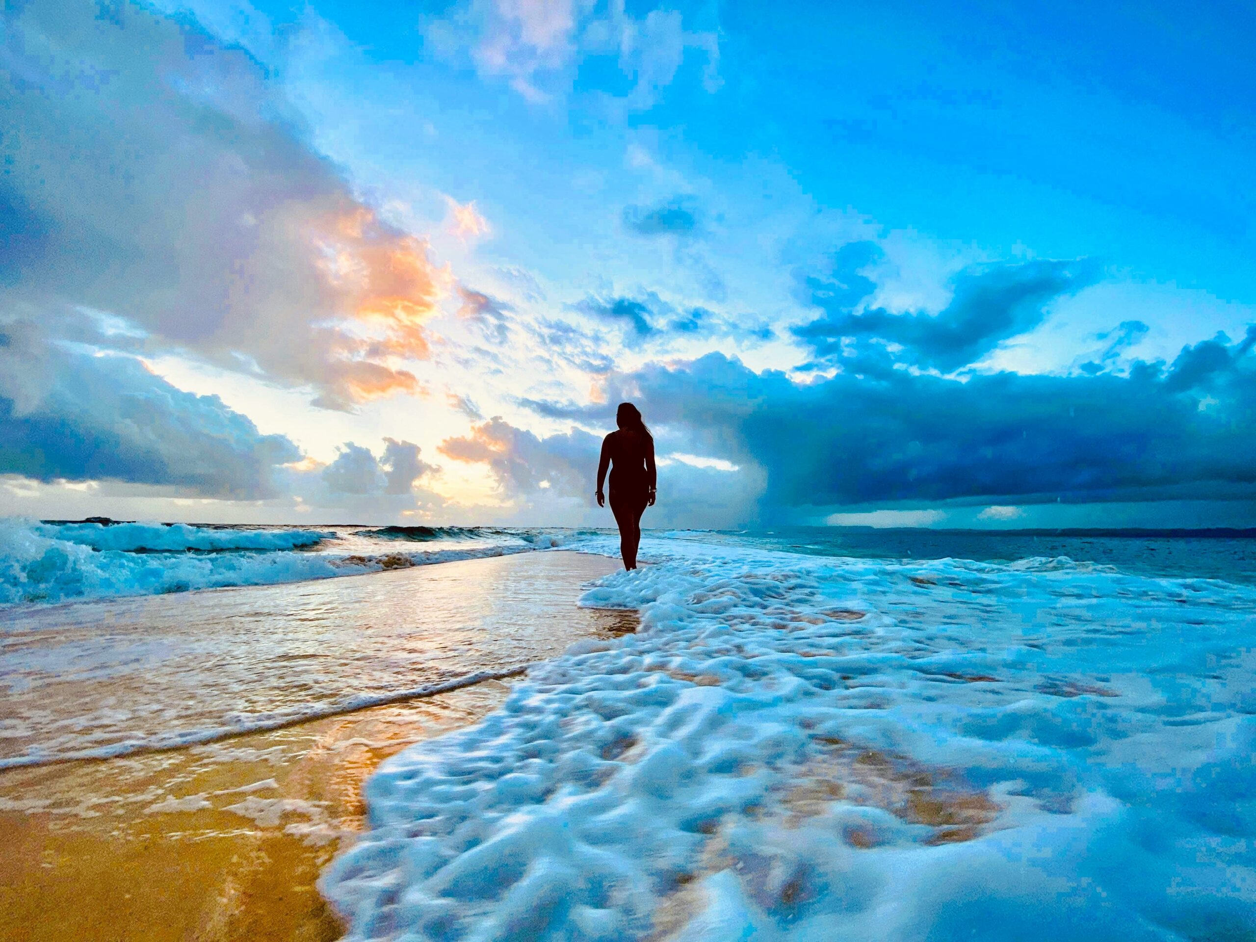 A lone figure walks barefoot along the shoreline beneath a vast blue sky